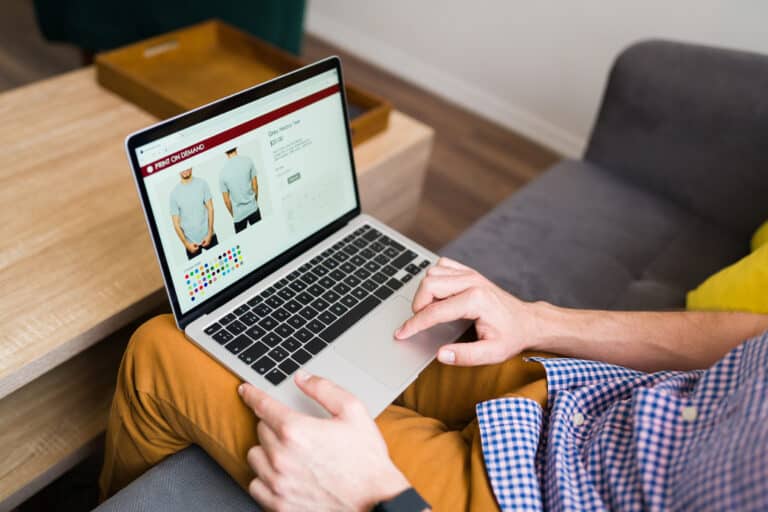 A person on a couch uses a laptop showing a print-on-demand t-shirt page with color options.