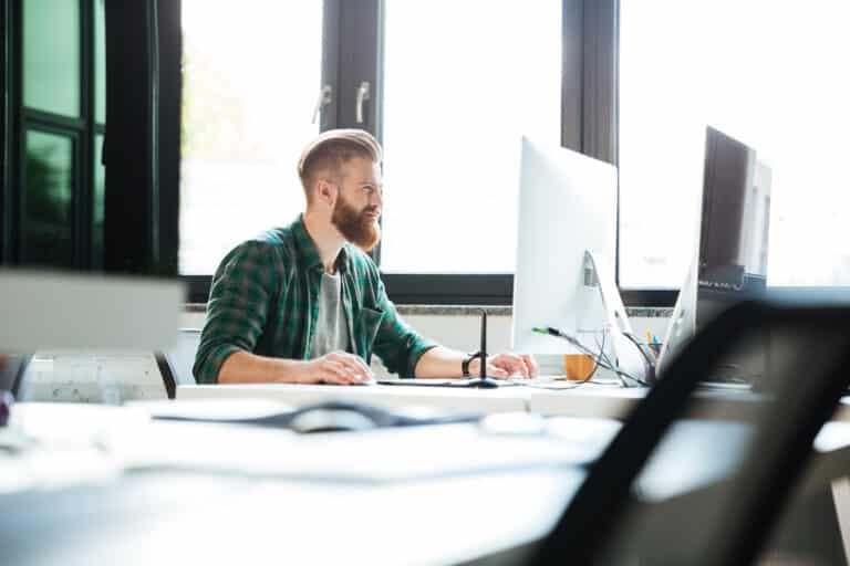A bearded man in a green plaid shirt works at a bright office desk with two monitors.