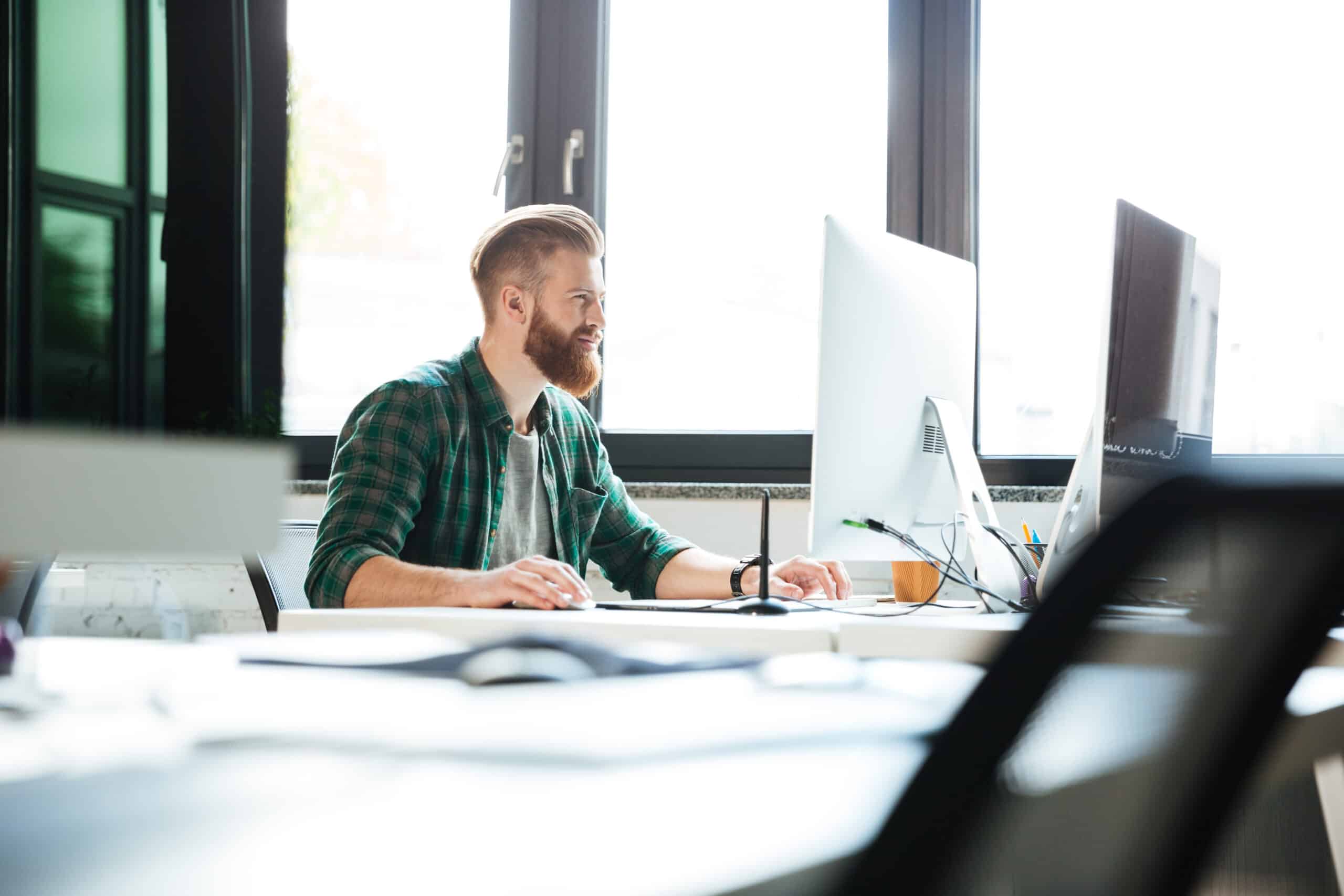 A bearded man in a green plaid shirt works at a bright office desk with two monitors.
