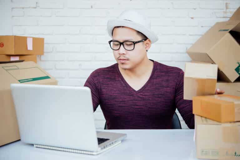 A man in a white hat and glasses works on a laptop beside stacks of shipping boxes.