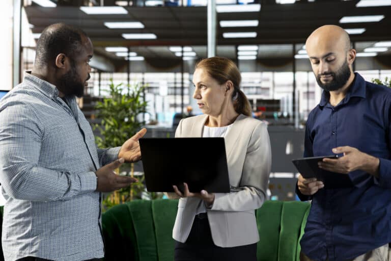 Three coworkers stand in an office; one holds a laptop while another looks at a tablet.