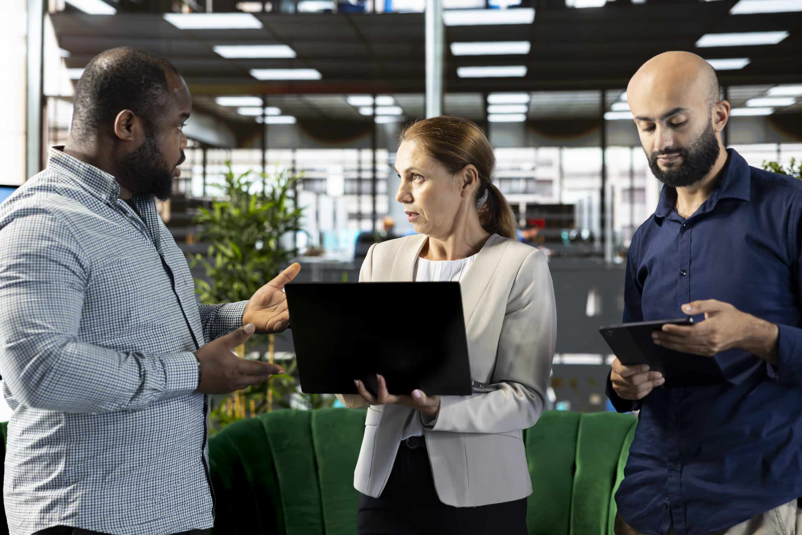 Three coworkers stand in an office; one holds a laptop while another looks at a tablet.