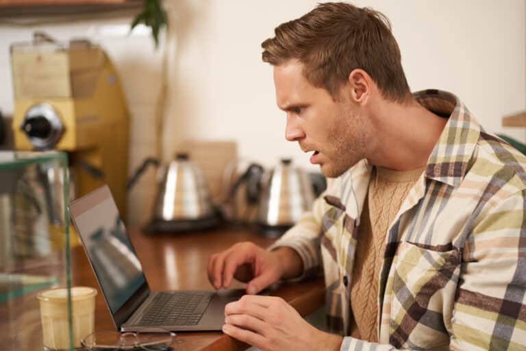 A man in a plaid shirt stares at a laptop in a cafe with a surprised, frustrated expression.