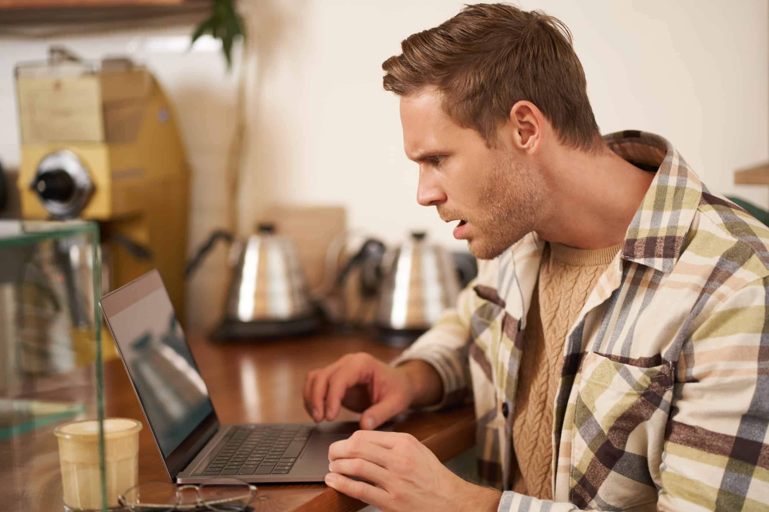 A man in a plaid shirt stares at a laptop in a cafe with a surprised, frustrated expression.