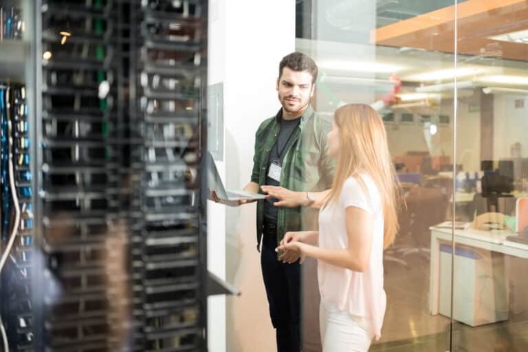 A man holding a laptop talks with a woman next to a glass-walled server rack.
