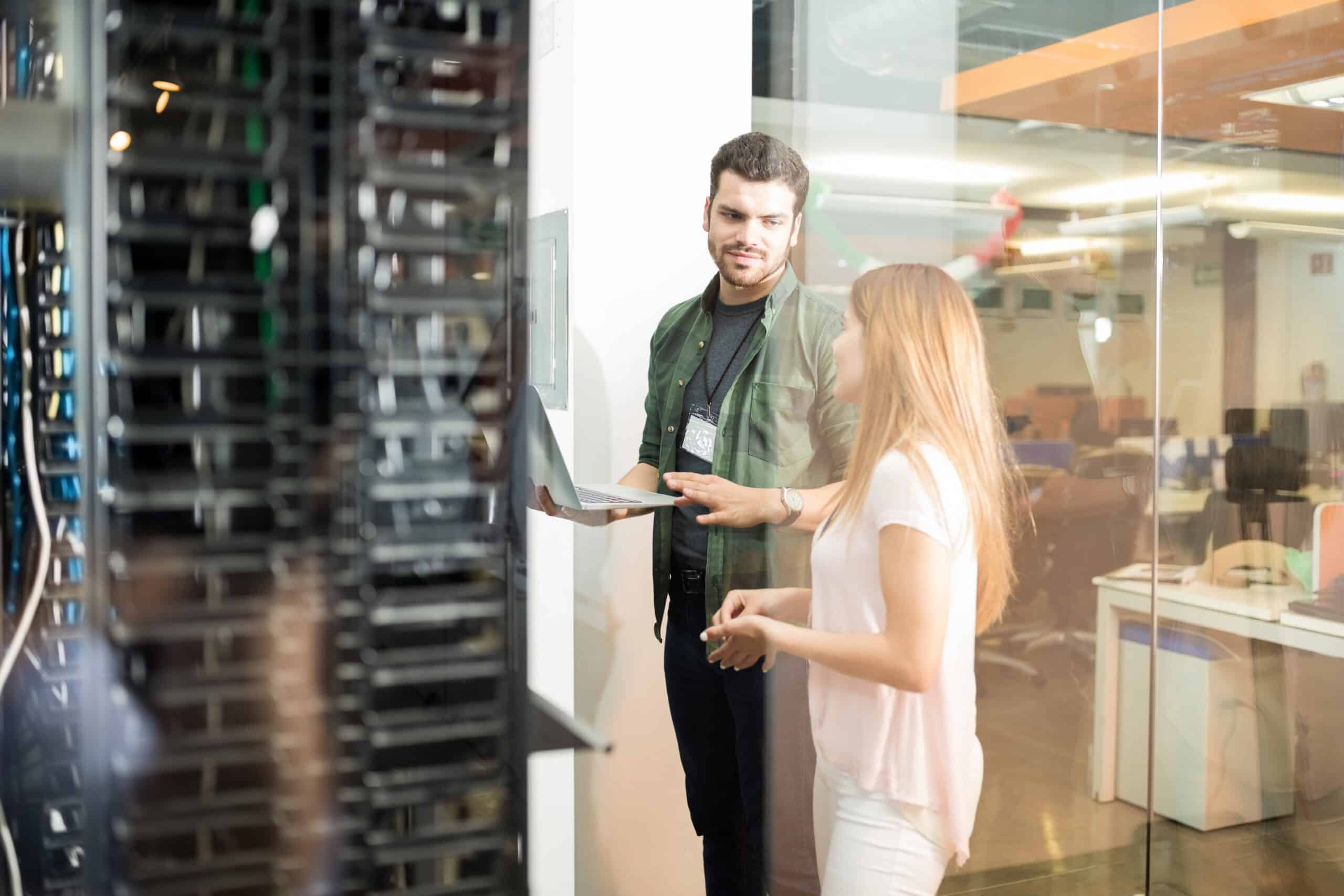 A man holding a laptop talks with a woman next to a glass-walled server rack.