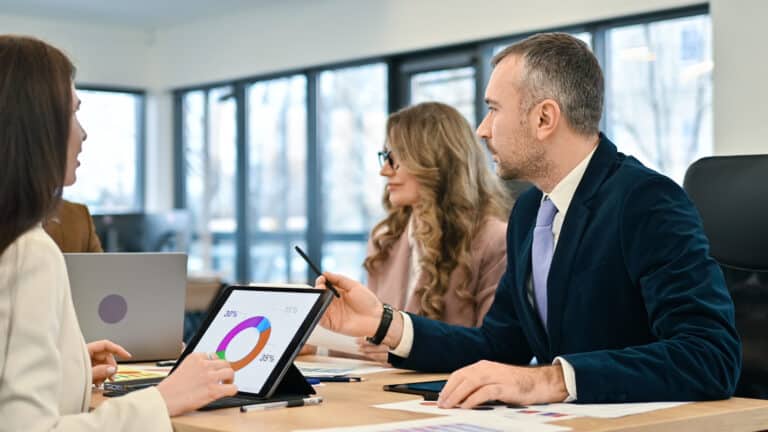 Three colleagues in a glass-walled office discuss a donut chart shown on a tablet.