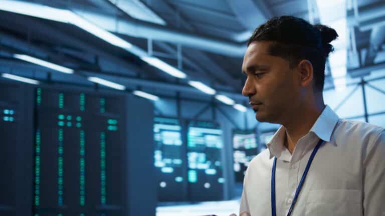 A man wearing a lanyard stands in a blue-lit data center beside racks of servers.