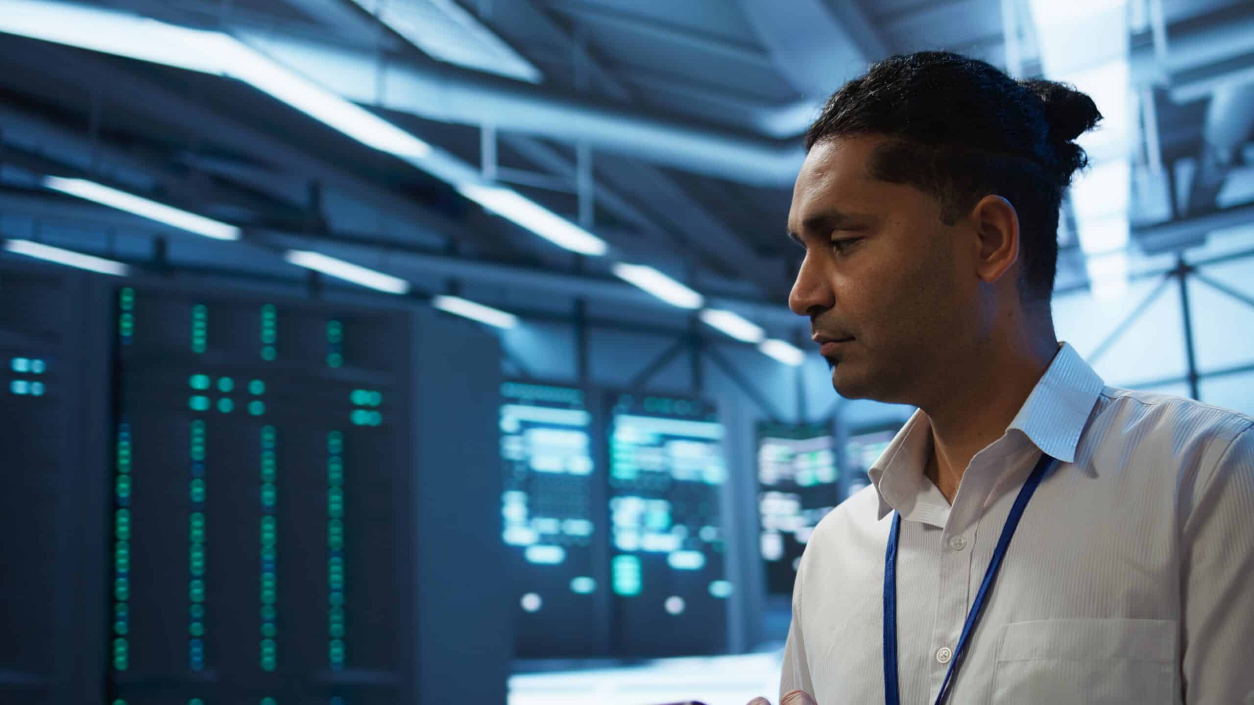 A man wearing a lanyard stands in a blue-lit data center beside racks of servers.